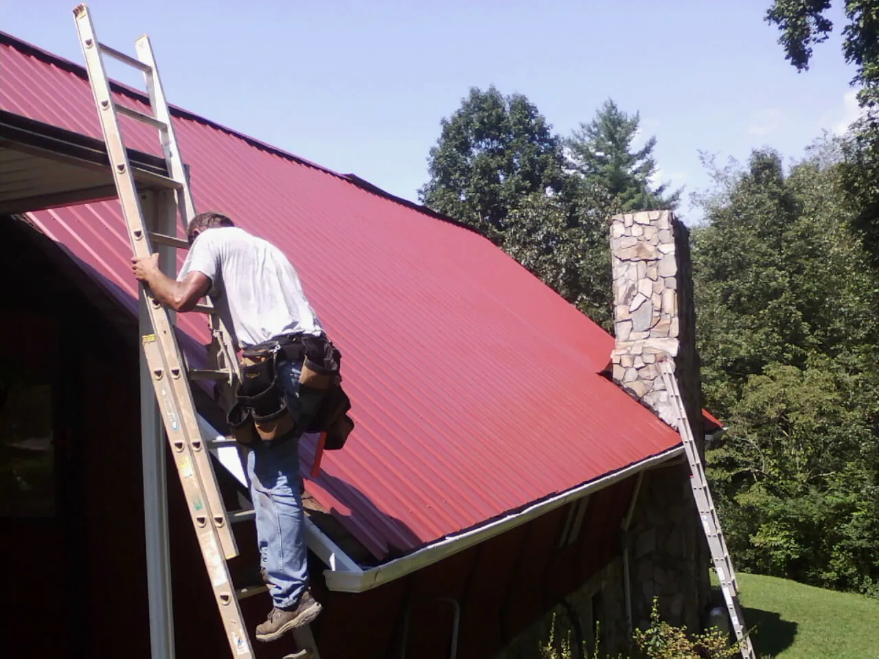 Roofer on a ladder beside a red metal roof and stone chimney — Soffit & Fascia Repair by Roofing Brothers in Midlothian